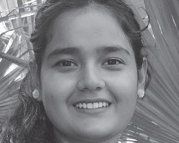 A woman with long hair smiles at the camera. She is wearing earrings and a background of palm leaves is visible.